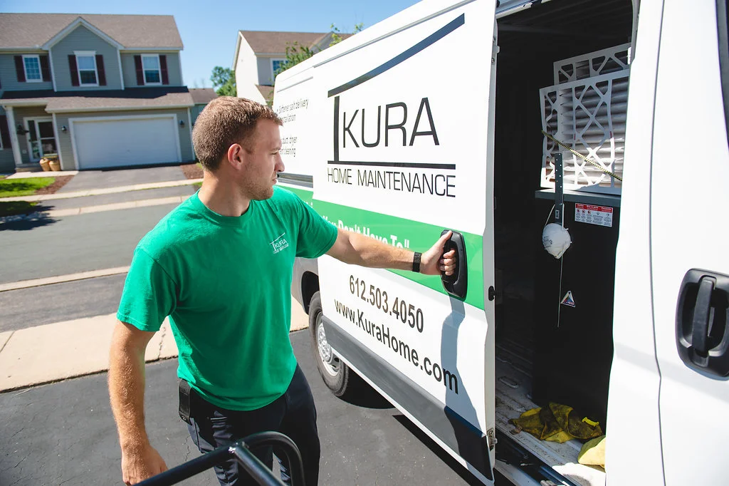 Technician in green shirt opening Kura Home service van on suburban street with houses in background and equipment visible inside the van.