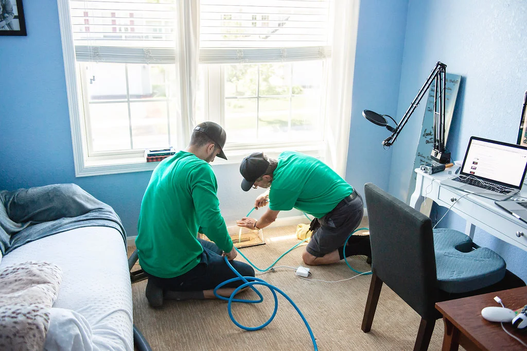 Technicians in green uniforms cleaning floor vent with blue air hoses in bedroom next to window bed chair and desk.