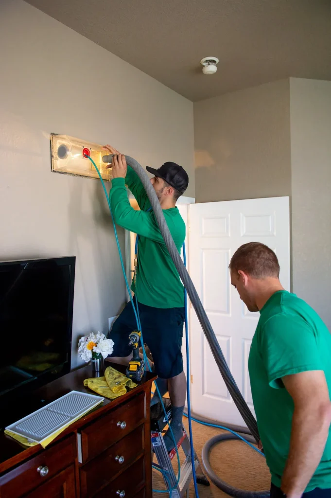 Technicians in green uniforms cleaning wall vent using clear vacuum seal and air hose with ladder drill and supplies on dresser in bedroom.